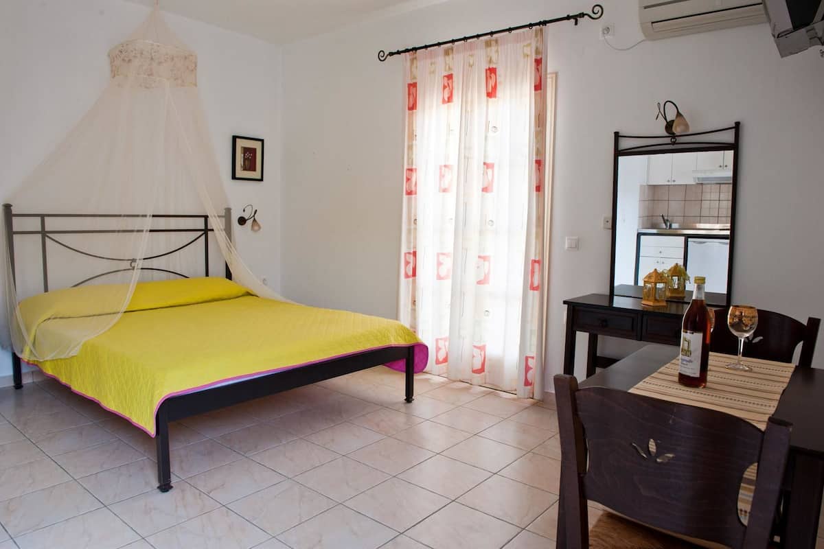 A bedroom with a bed, a desk, a chair, a mirror, a window with curtains, and an air conditioning unit.