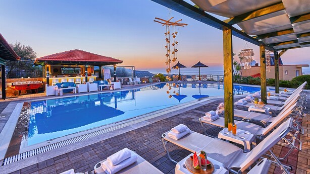 A poolside area with lounge chairs, a bar, and a view of the sea at sunset. - Everest Apartments (Hersonissos)