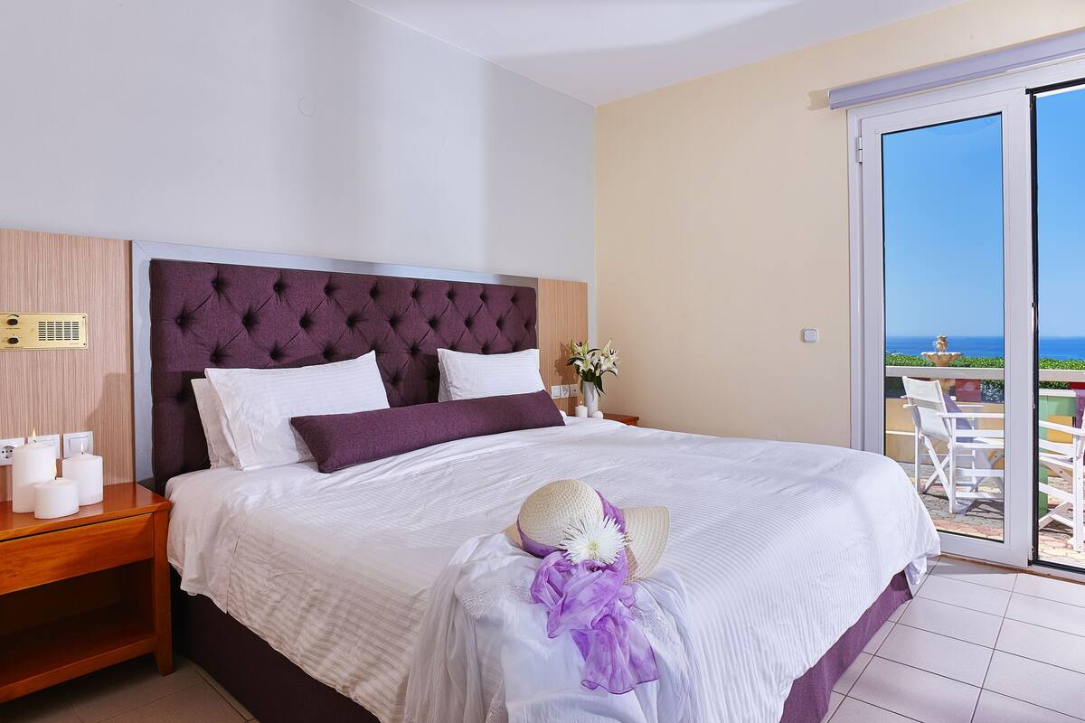 A hotel room with a large bed, a wooden nightstand, a white wall, and a view of the ocean through a sliding glass door.