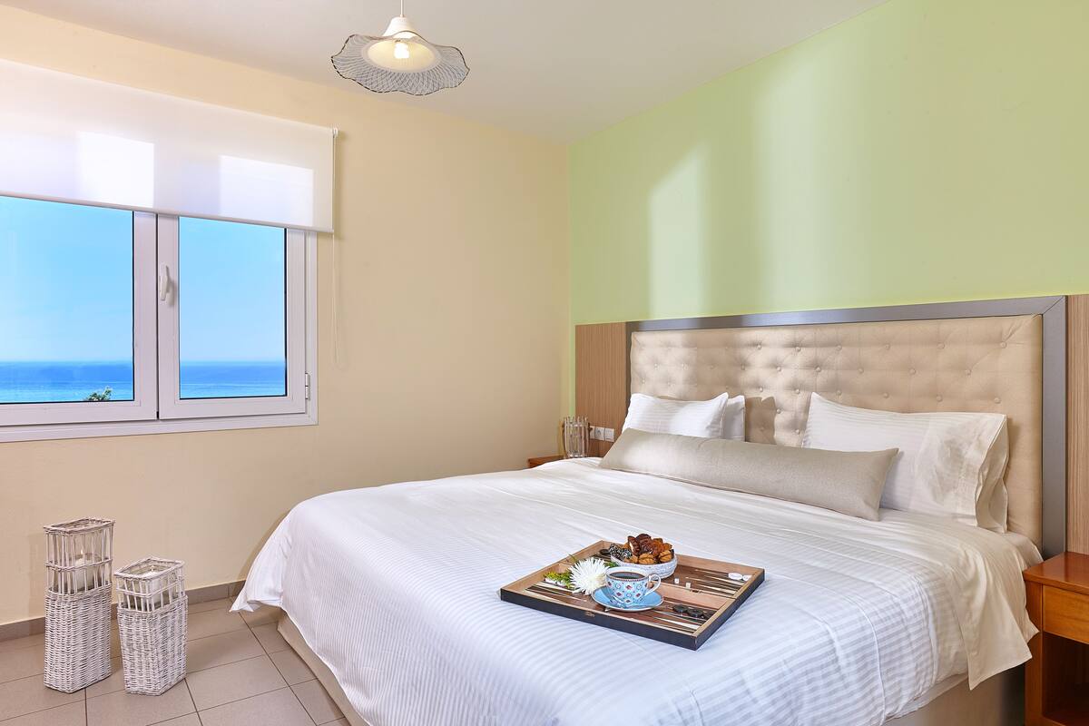 A neatly made bed with a tray of breakfast items, a view of the sea through the window, and a simple, clean room design.