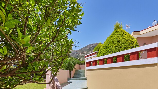 A staircase leading up to a building with a beige exterior and red accents, surrounded by greenery and a clear blue sky. - Everest Apartments (Hersonissos)