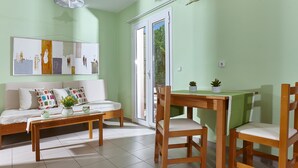 A living room with a sofa, a wooden coffee table, and two wooden chairs. There is a dining table with a green tablecloth and a small potted plant on it. - Everest Apartments (Hersonissos)