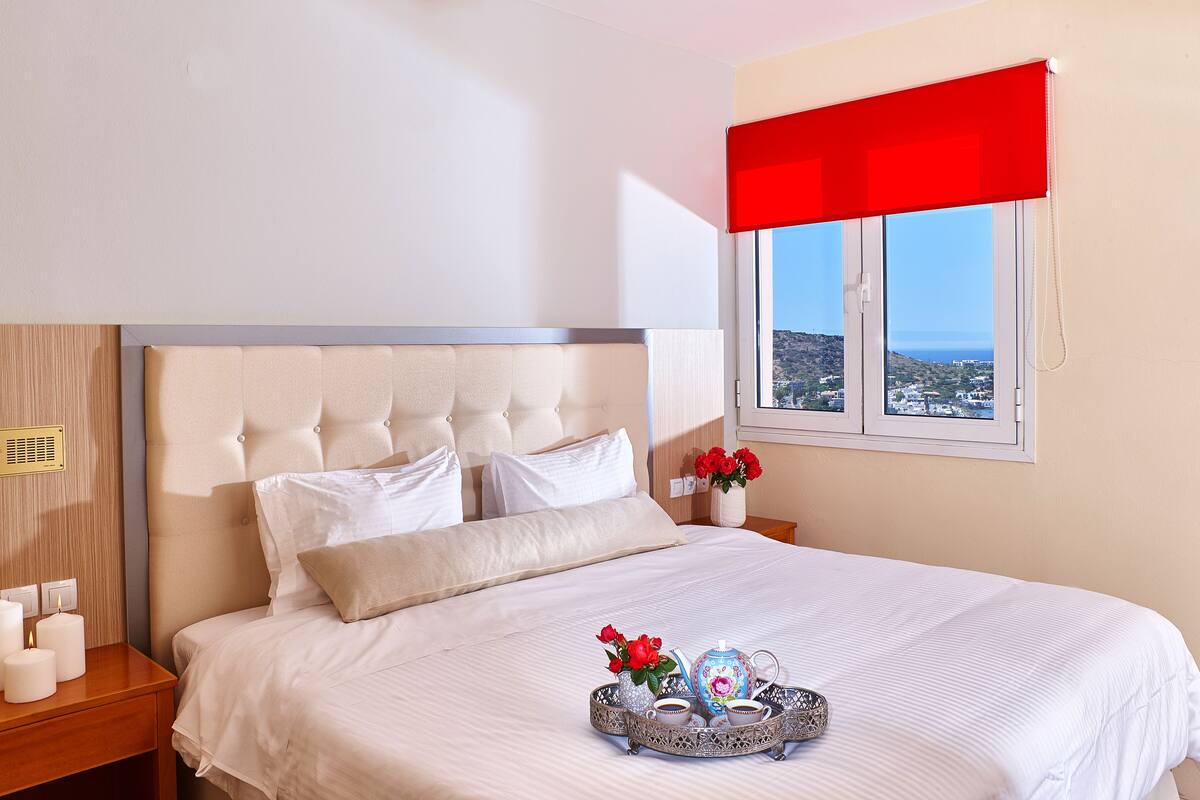 A neatly made bed with white linens, a bedside table with candles, and a tray with a teapot and flowers.