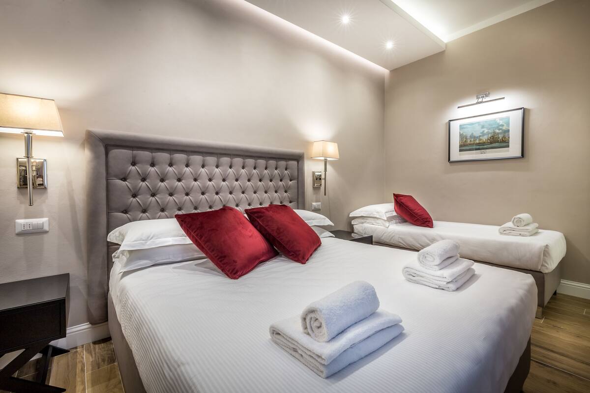 A hotel room with a large bed, two smaller beds, red and white pillows, a framed picture on the wall, and a lamp on the nightstand.