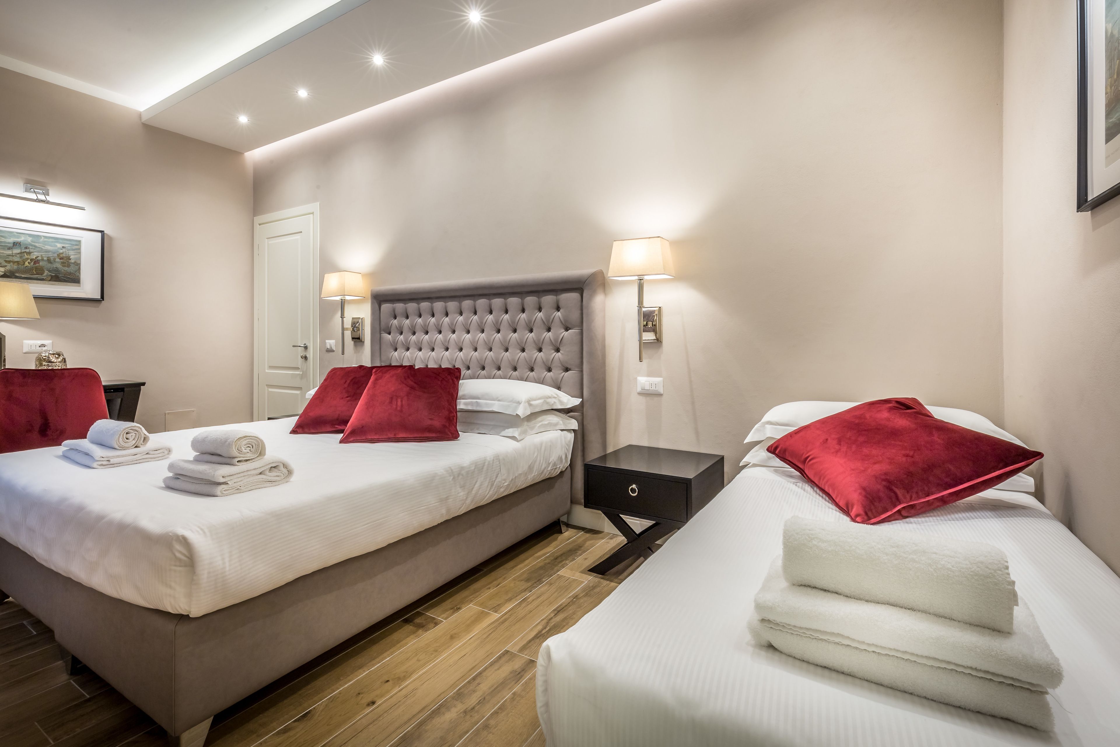 Une chambre d’hôtel avec deux lits, chacun agrémenté d’un oreiller rouge et de draps blancs. Il y a deux tables de chevet avec des lampes et un petit tiroir. La chambre est décorée dans des tons neutres, avec des murs de couleur beige et un plancher en bois.