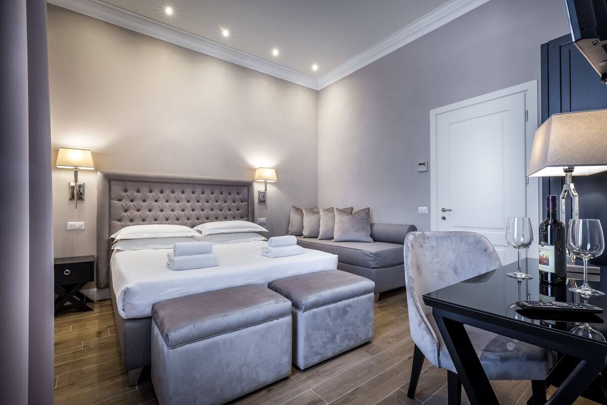 A modern hotel room with a large bed, a sofa, a dining table, and a minibar.