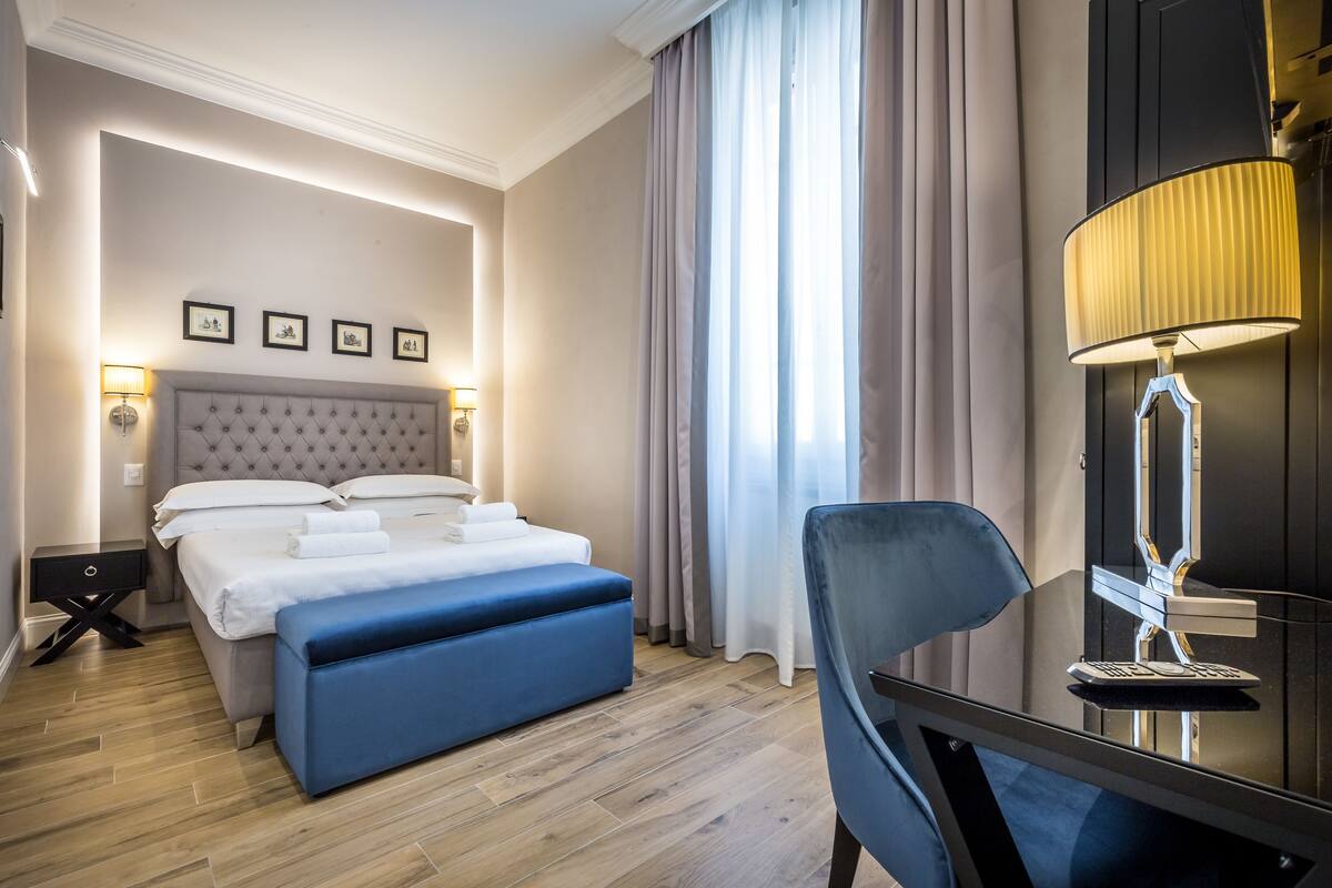A hotel room with a bed, a desk, a chair, a lamp, and a window with curtains.