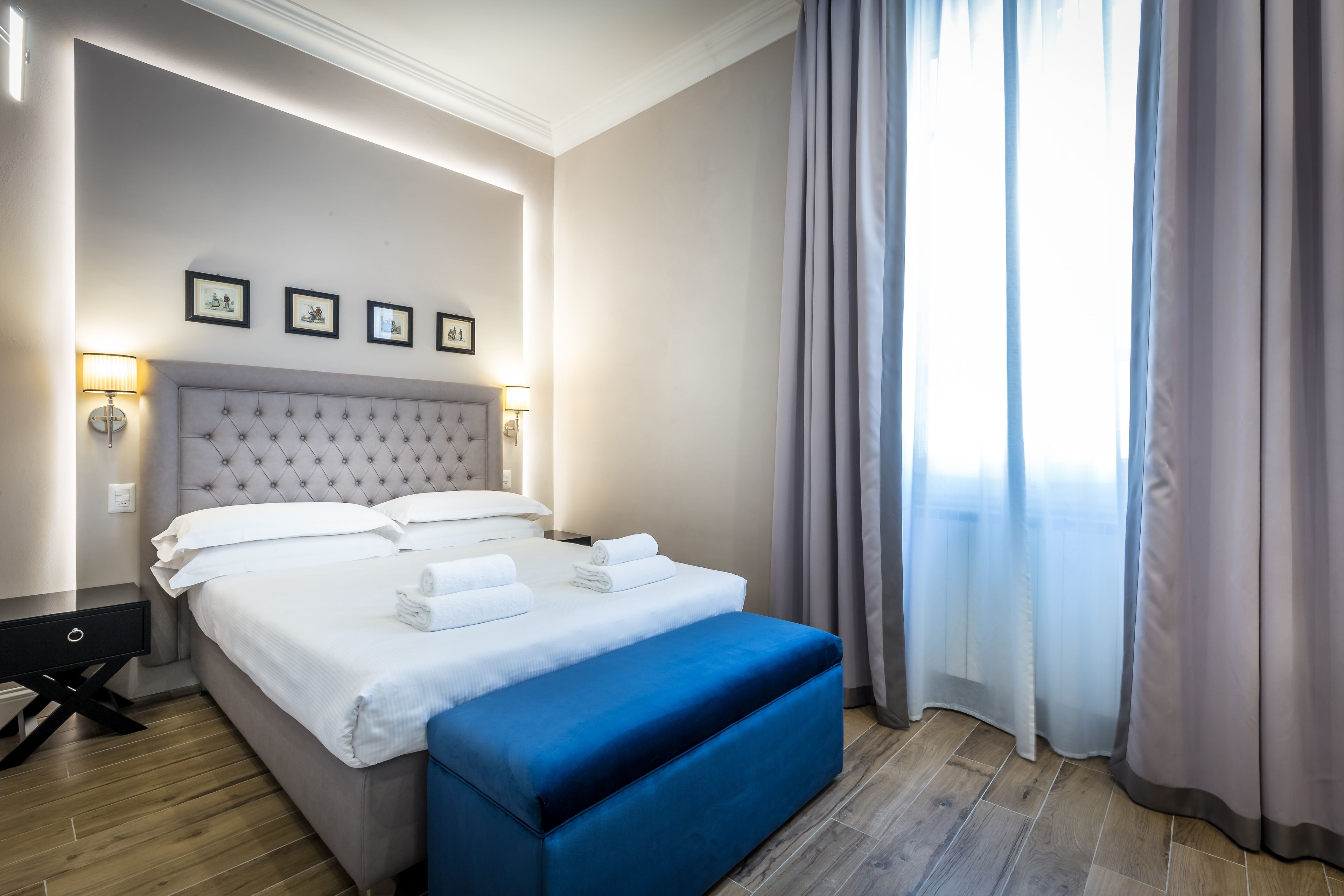 Una cama bien hecha con ropa de cama blanca y un banco azul, una mesita de noche con lámpara, y una pared con cuadros enmarcados.