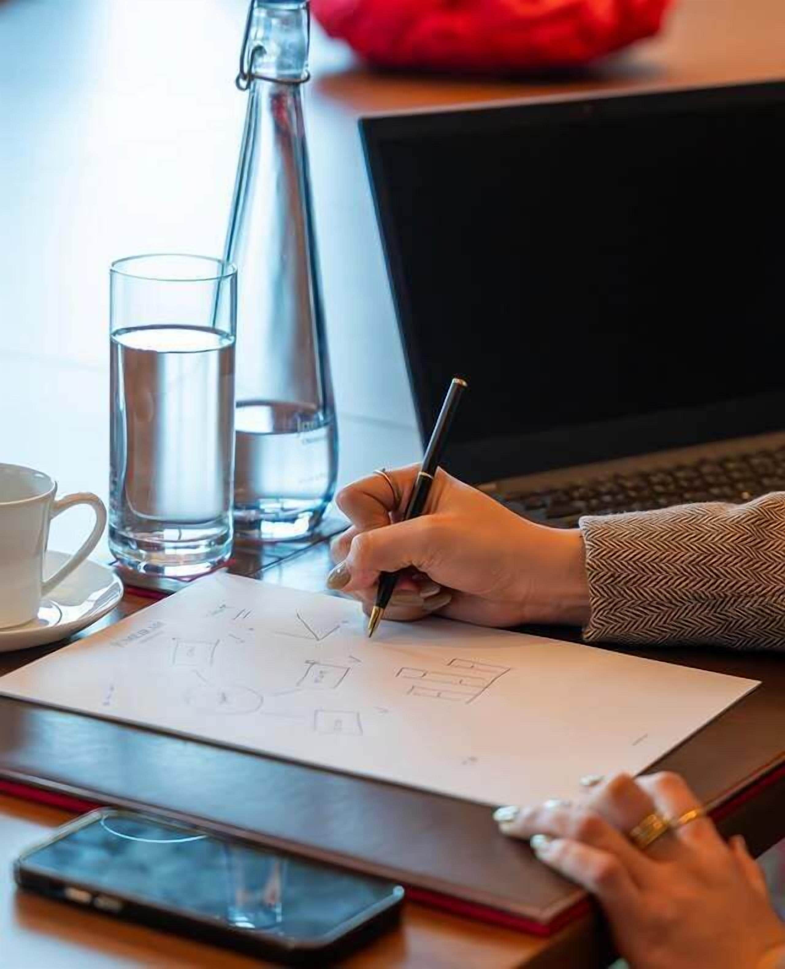 A person writing on a paper with a pen, a laptop, a glass of water, and a cup on a table.