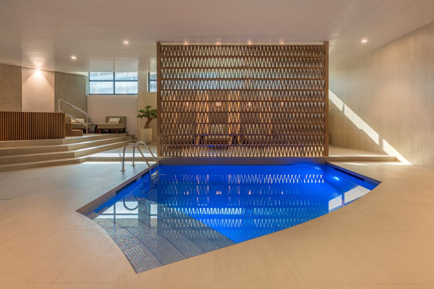A modern indoor pool with a geometric wall and a staircase leading to a seating area.