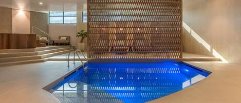 A modern indoor pool with a geometric wall and a staircase leading to a seating area.