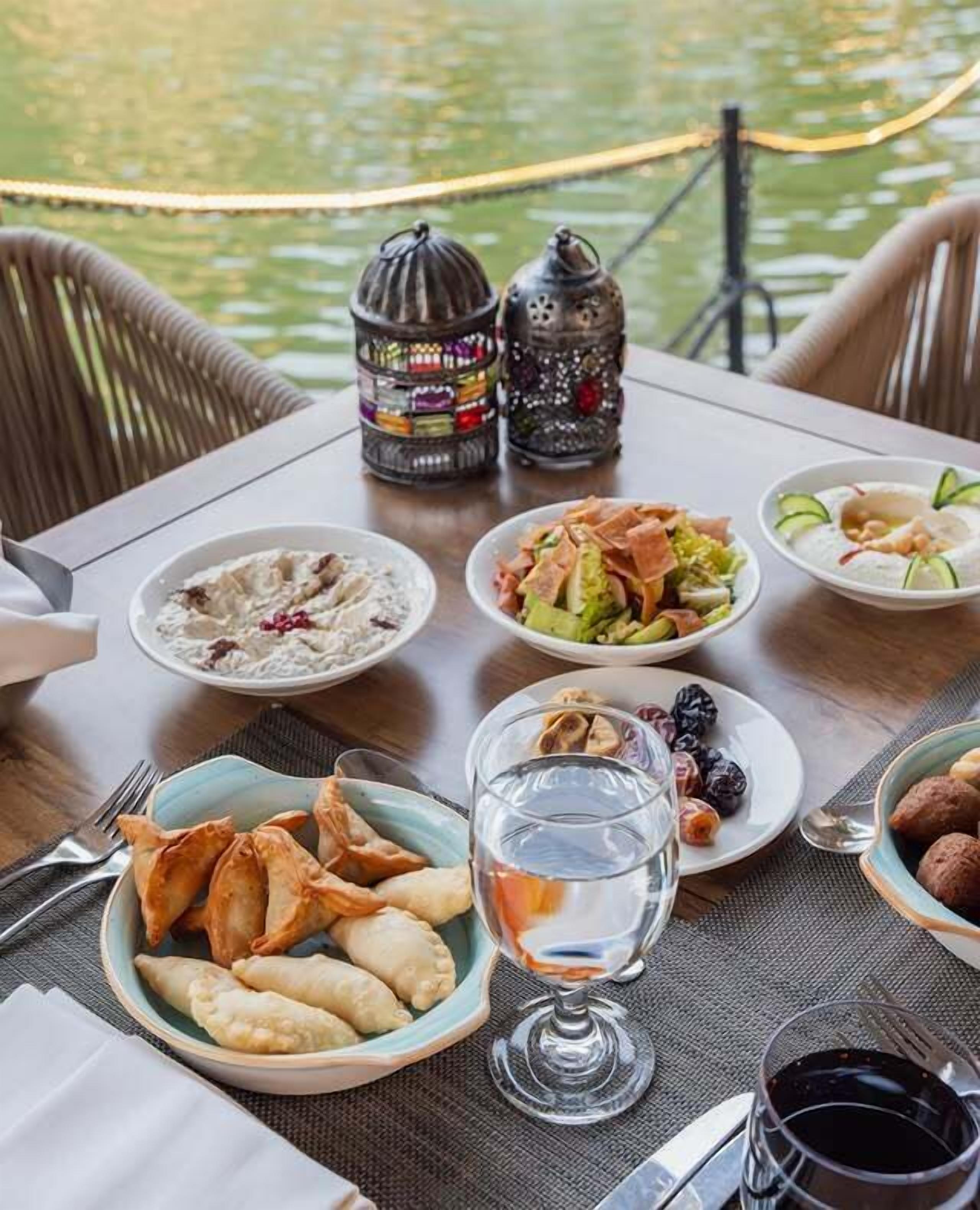 Una mesa dispuesta para una comida con diversos platos, incluyendo samosas, una ensalada y un cuenco de yogur con pepino. También hay dos faroles decorativos y una vista de una masa de agua al fondo.