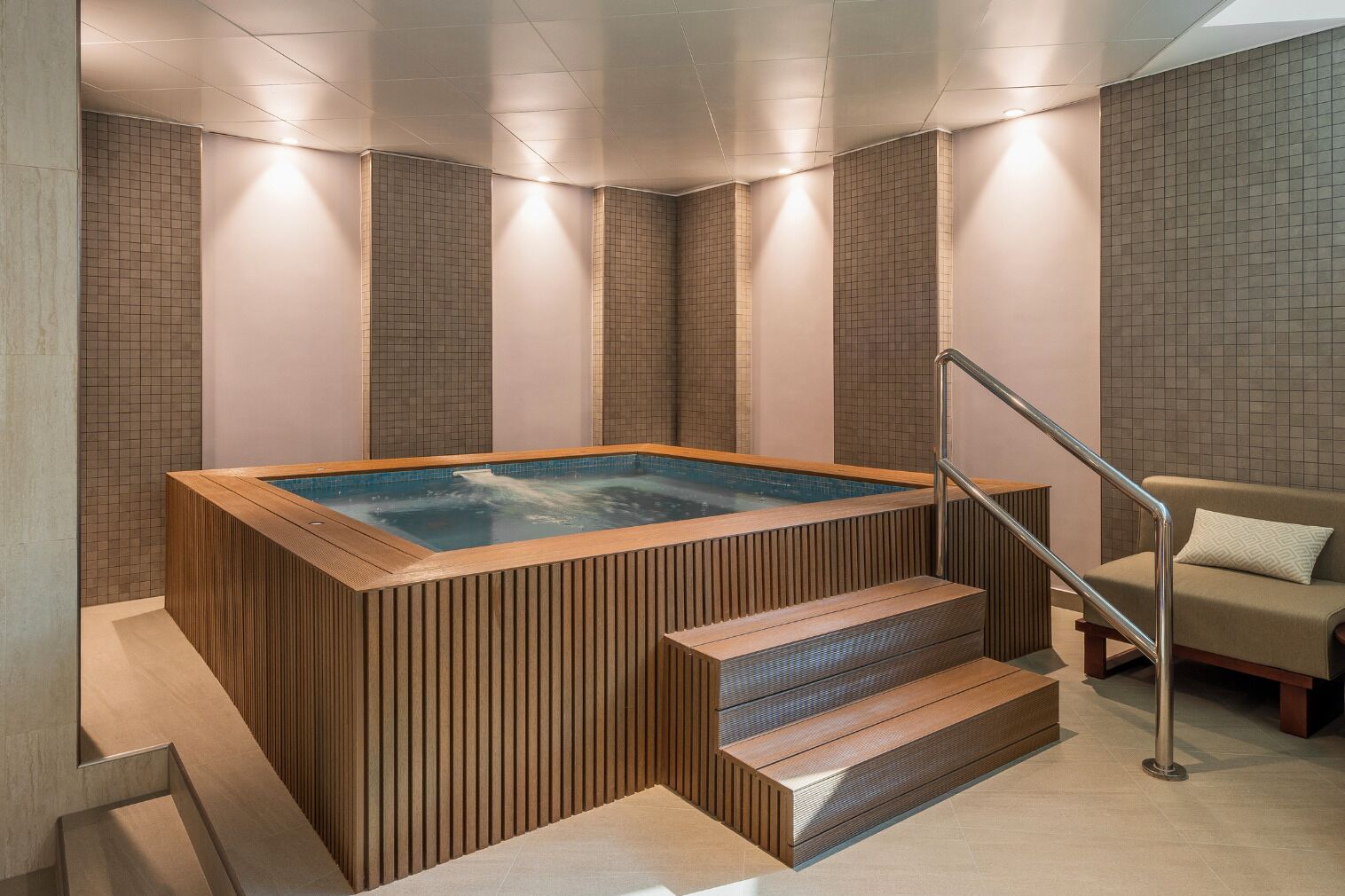 A modern bathroom with a wooden bathtub, a beige sofa, and a staircase leading to a higher level.