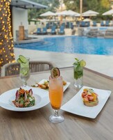 Una mesa con tres platos de comida y un vaso alto de bebida, frente a una vista de piscina con sombrillas y sillas de descanso.