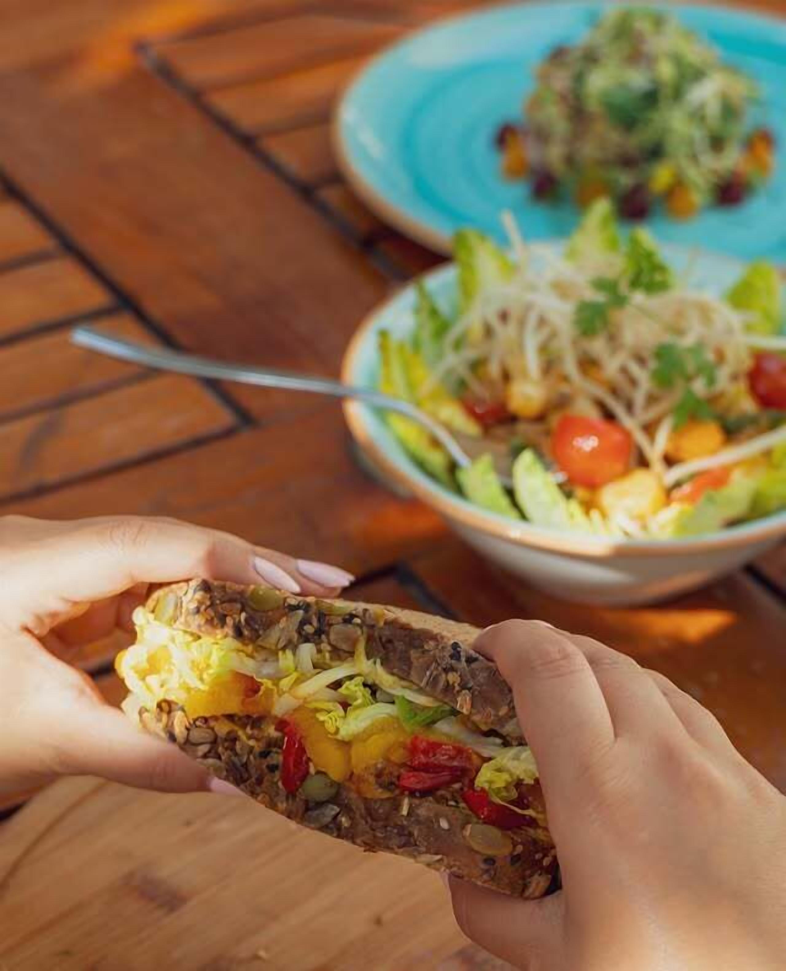 A person holding a sandwich with visible ingredients like lettuce, tomato, and possibly cheese. In the background, there is a bowl with a salad containing lettuce, tomatoes, and other vegetables.