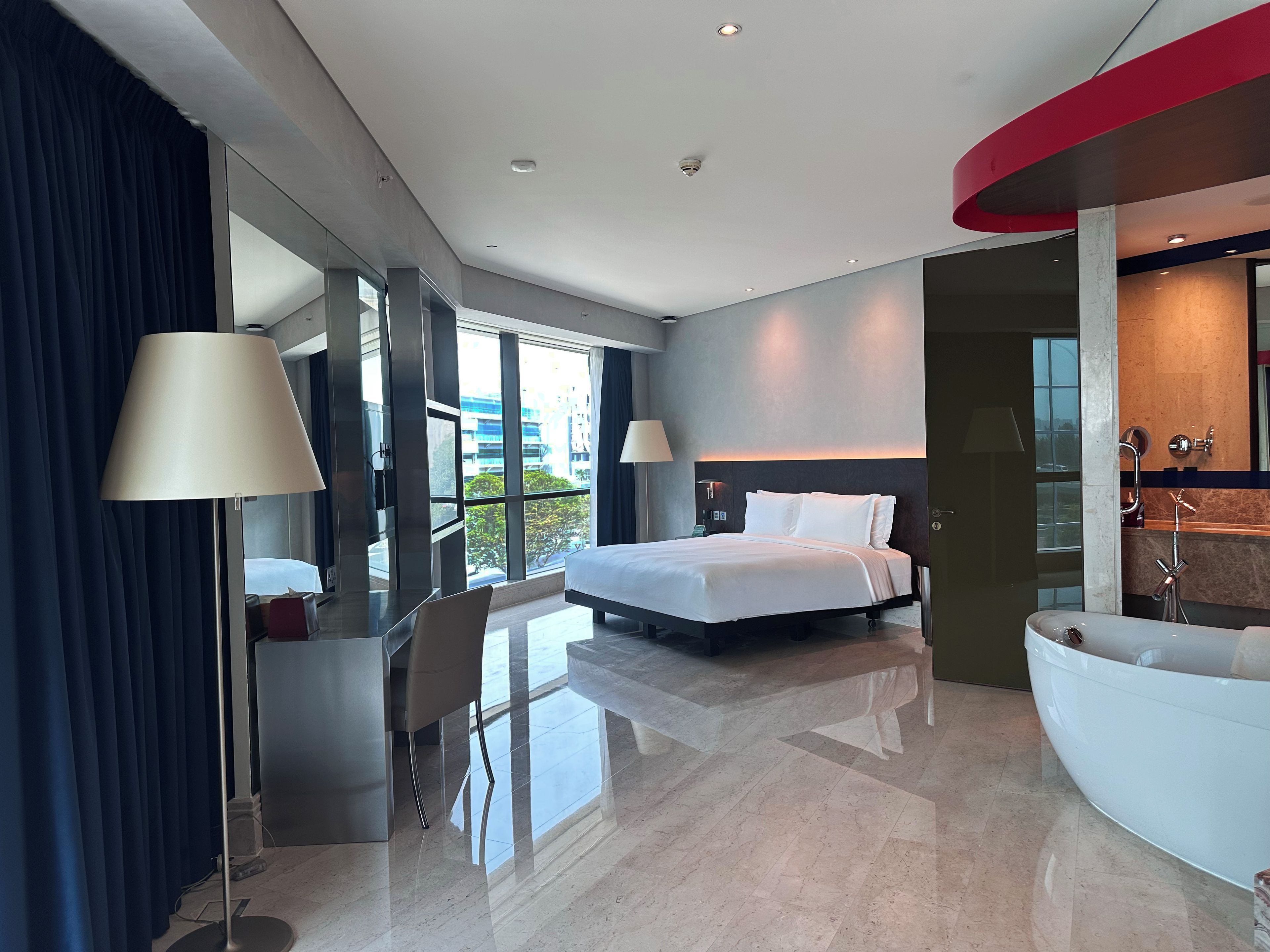 A modern hotel room with a large bed, a sitting area with a desk, a large window, and a separate bathroom with a bathtub.