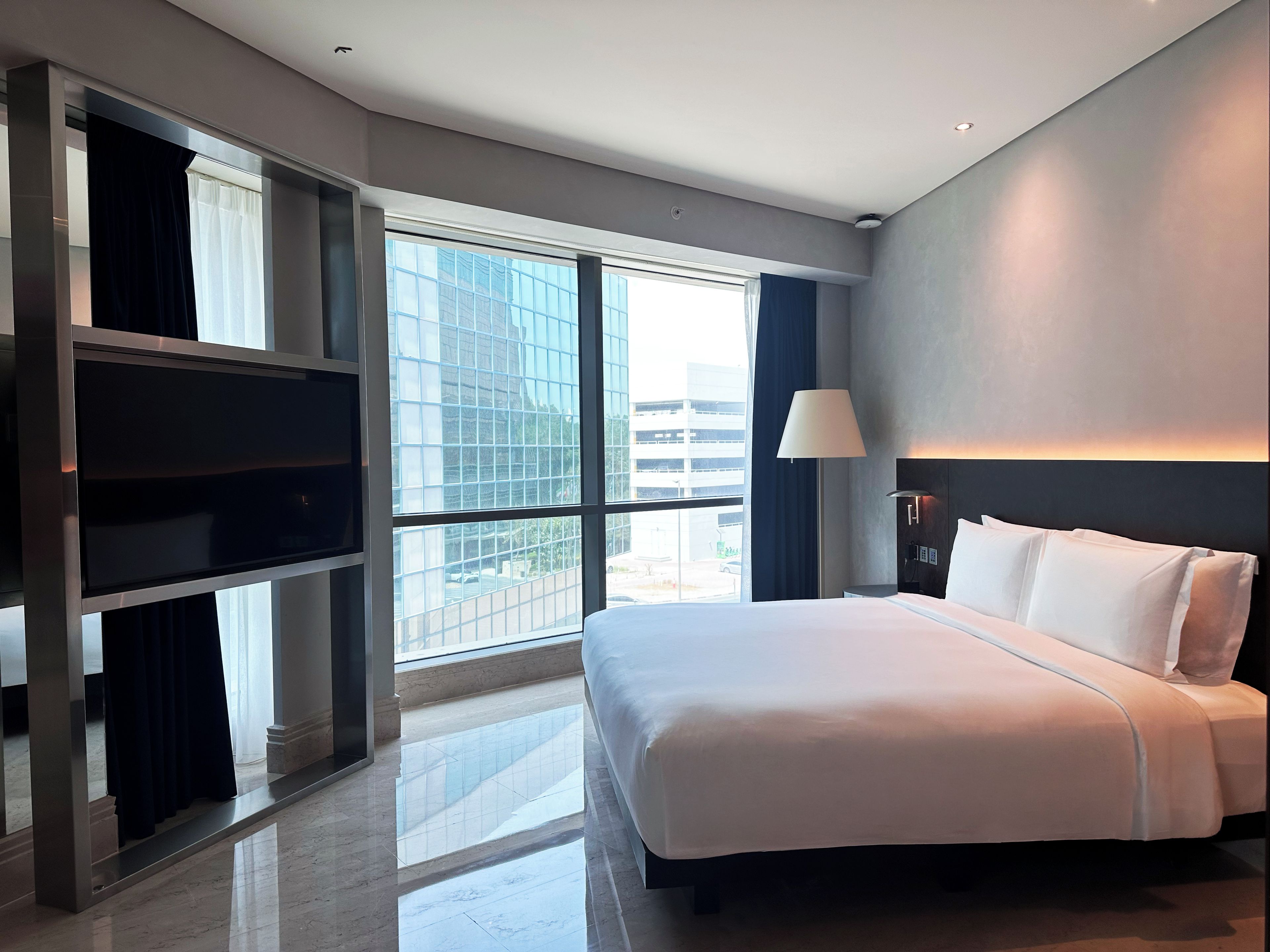 A modern hotel room with a large bed, a TV, and a view of the city through large windows.