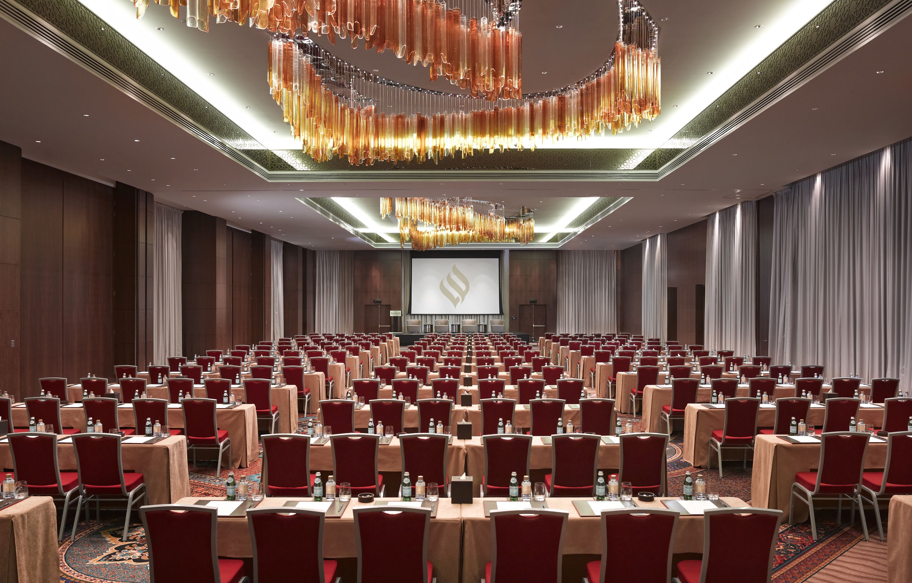 Une salle de conférence avec des rangées de tables et de chaises, un grand écran à l’avant et un lustre.