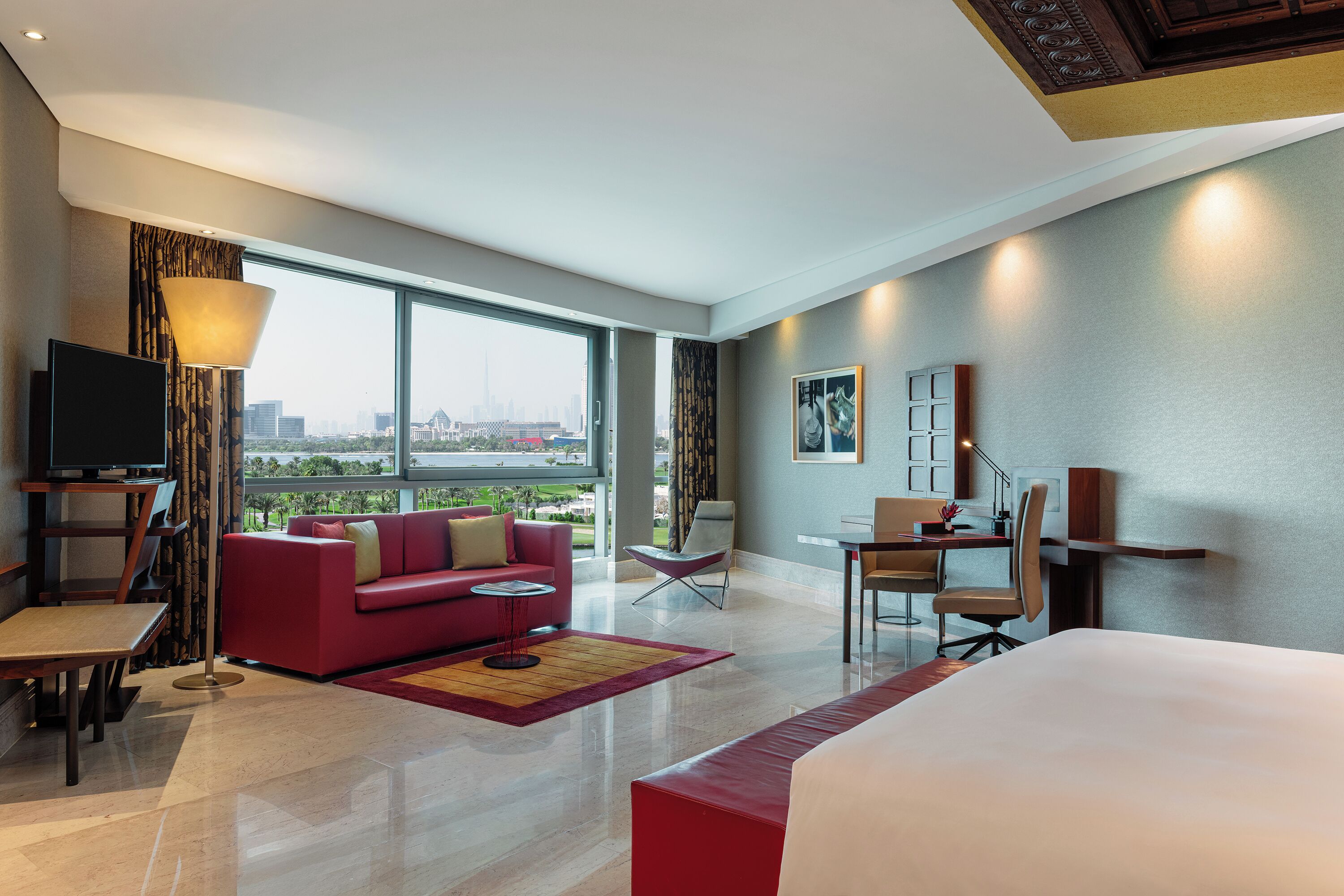 A modern hotel room with a large bed, a red sofa, a desk, a TV, and a city view through large windows.