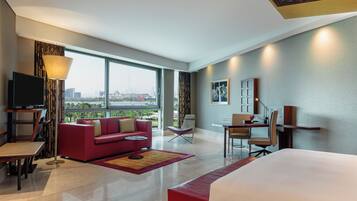 A modern hotel room with a large bed, a red sofa, a desk, a TV, and a city view through large windows.