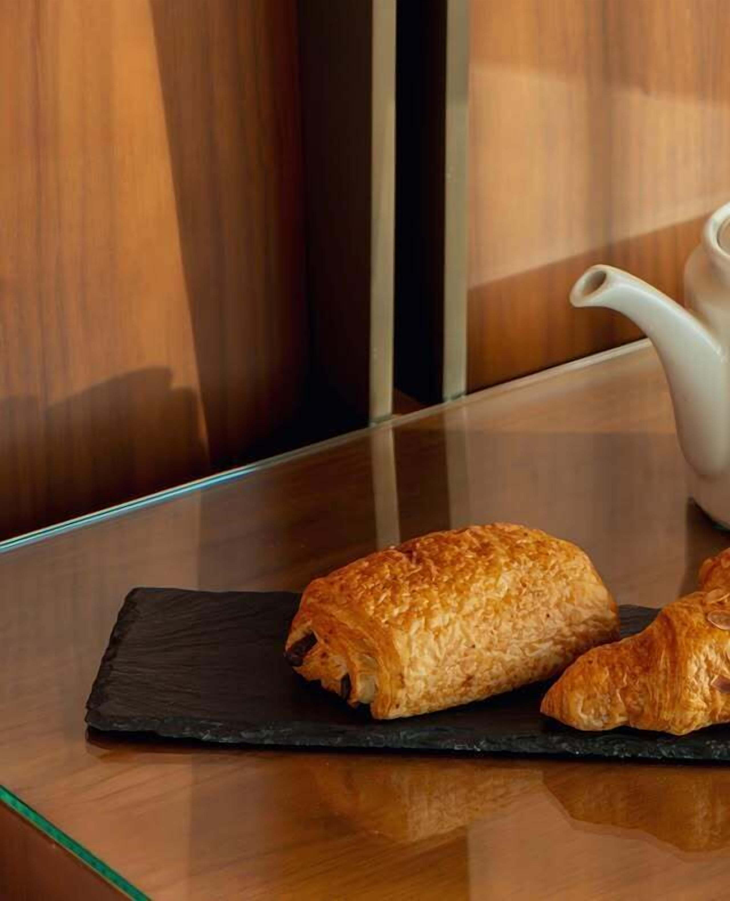 Deux pâtisseries sur une assiette en ardoise noire, posée sur une table en verre, avec une théière blanche en arrière-plan.