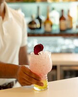 A person holding a pink cocktail with a rose garnish, with bottles in the background.