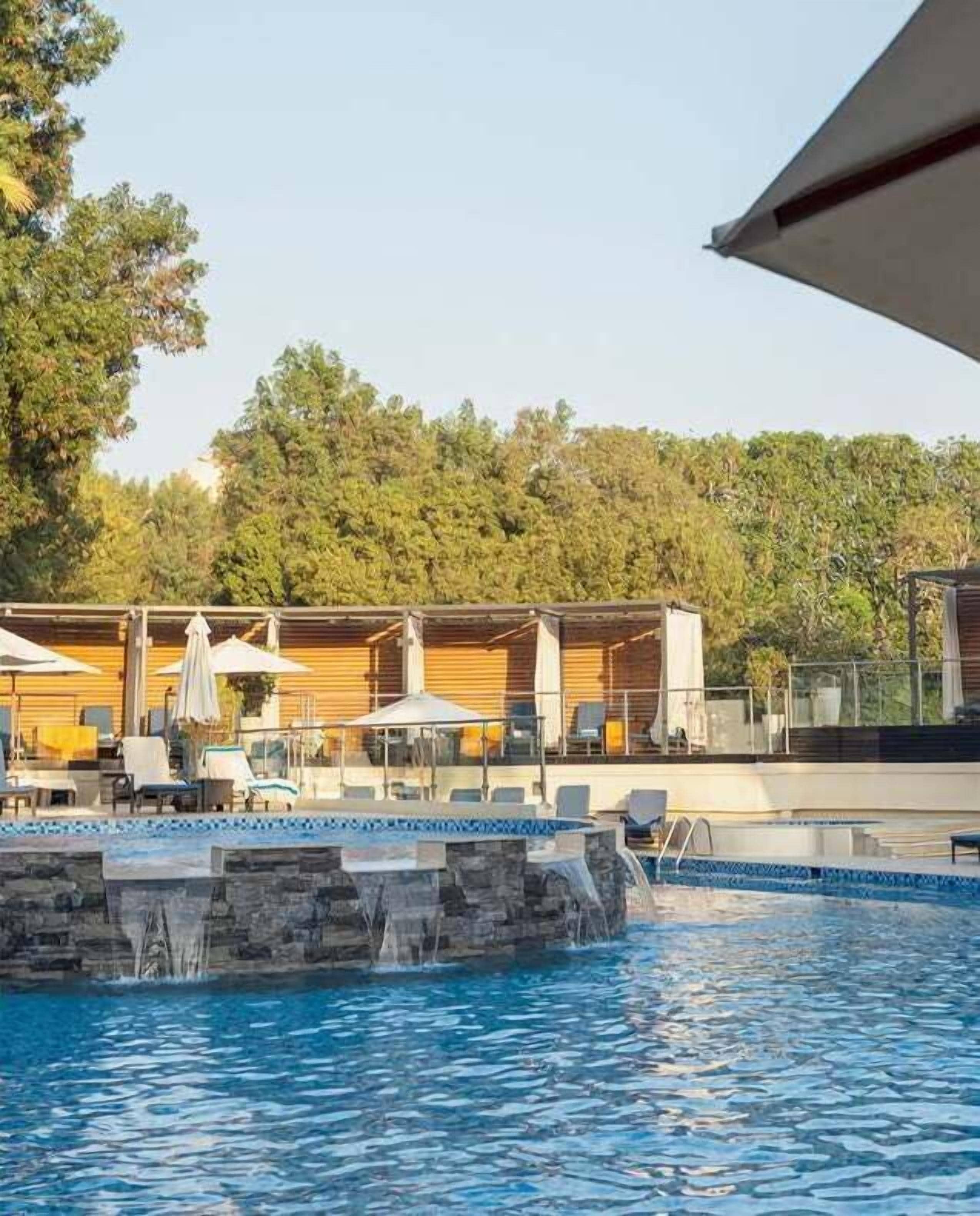 En pool med ett vattenfall, omgiven av solstolar och parasoller, och en träbyggnad syns i bakgrunden.