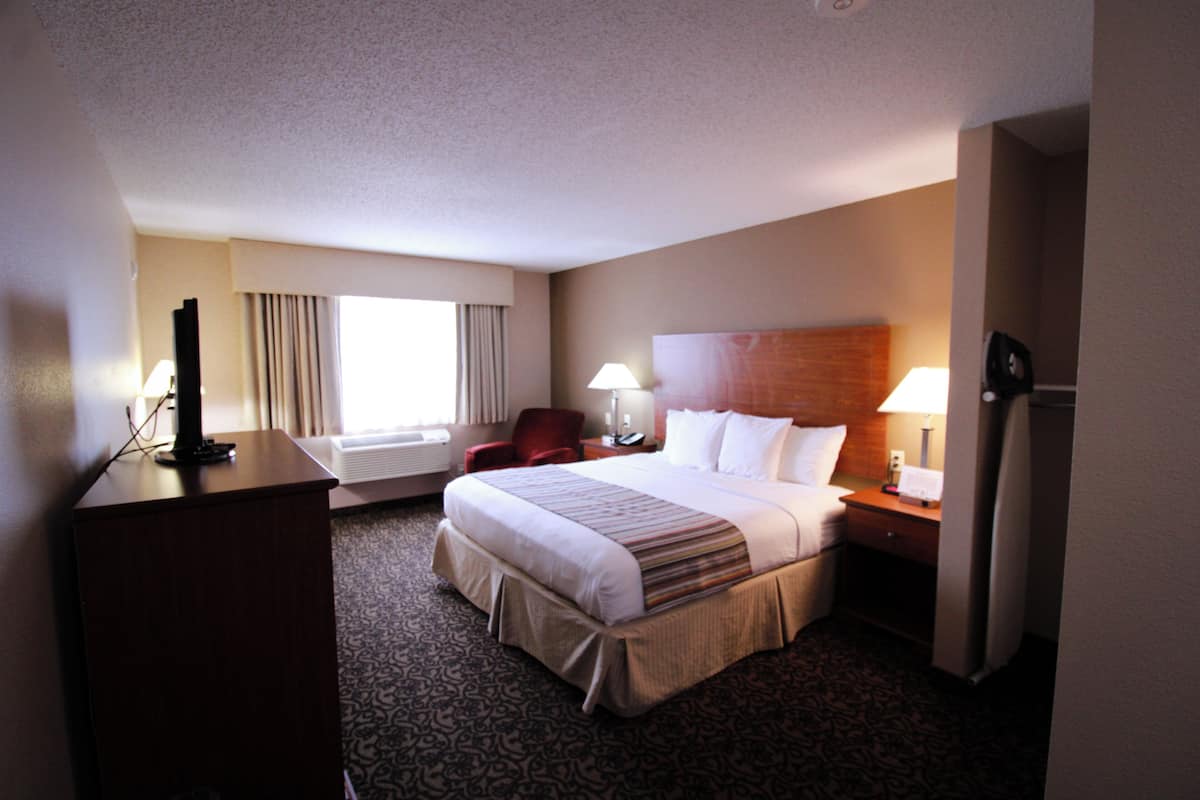 A hotel room with a large bed, a desk with a computer, a chair, a nightstand with a lamp, and a window with curtains.