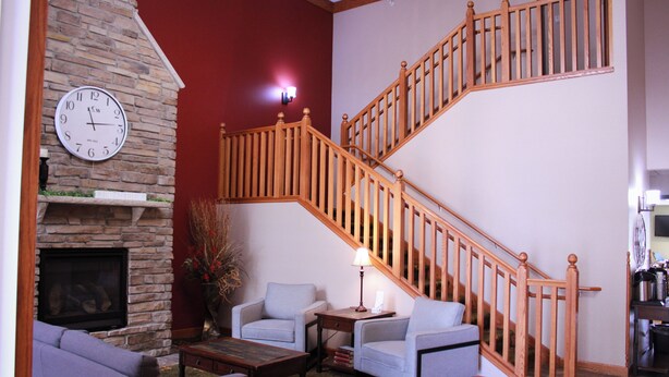 A living room with a fireplace, two armchairs, a coffee table, and a wooden staircase. - Canby Inn And Suites (Canby)