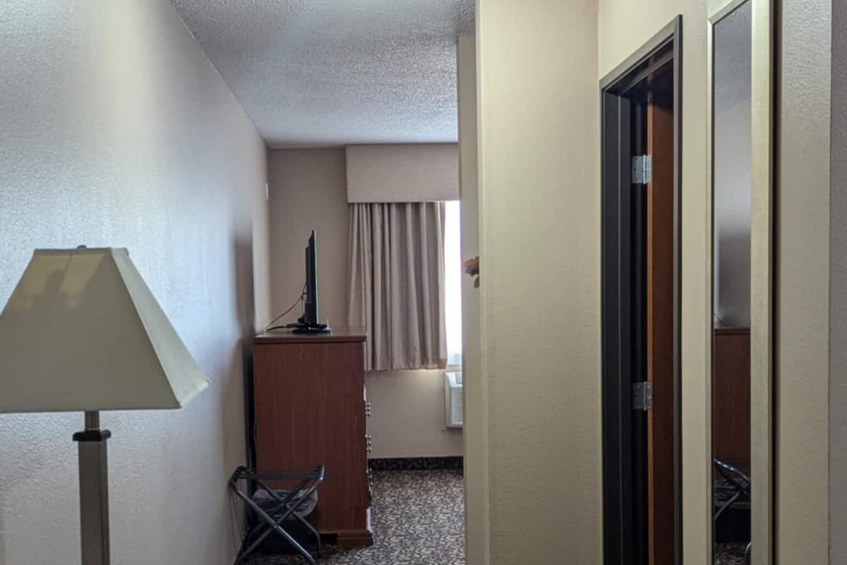 A hotel room with a desk, chair, lamp, and a small cabinet. There is a window with curtains and a door with a mirror.
