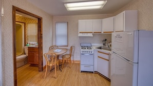 A compact kitchen with white cabinets, a stove, and a refrigerator. There is a dining table with chairs and a window with blinds. - Sea Spray Motel (Beach Haven)