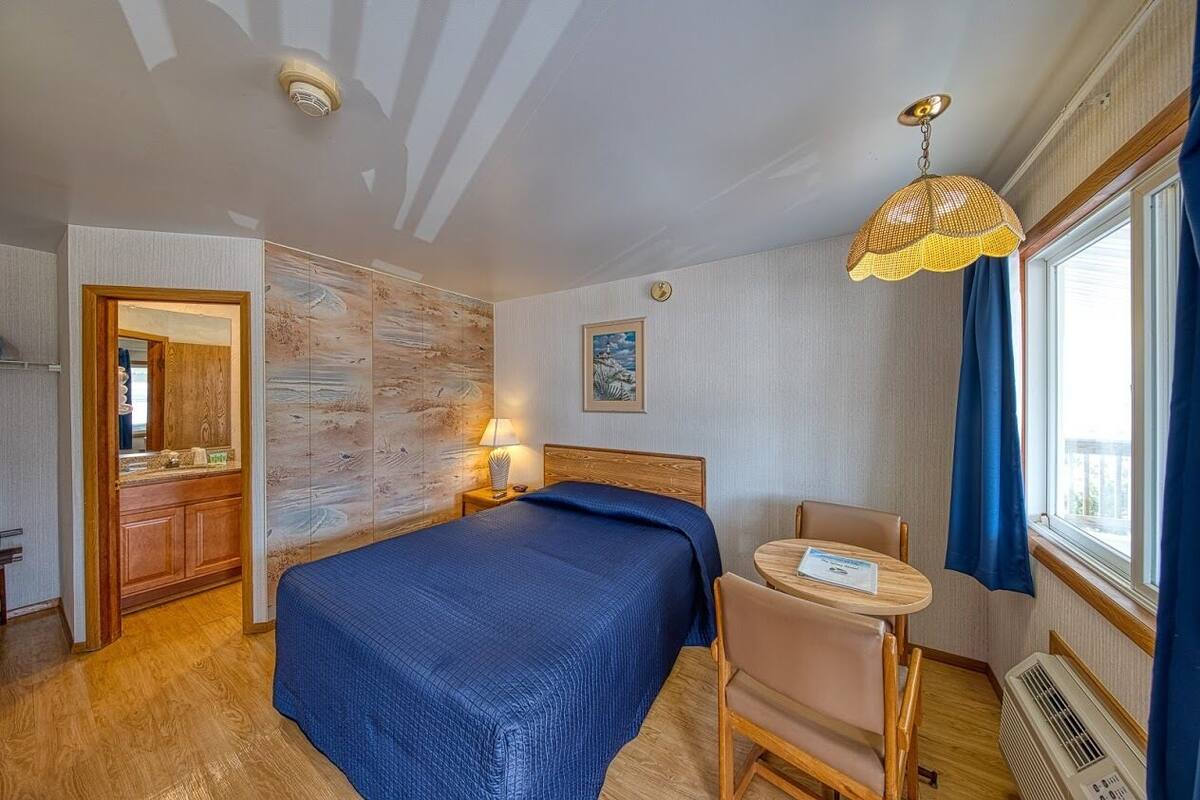 A hotel room with a bed, a desk, a chair, a lamp, a window with curtains, and a wall with a wooden panel.