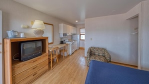 A compact room with a TV on a wooden stand, a small dining table with chairs, a sofa, and a kitchenette with cabinets and a microwave. - Sea Spray Motel (Beach Haven)