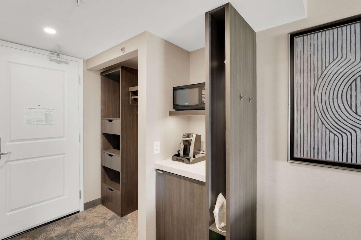 A modern room with a white door, built-in wooden shelving, a microwave, a coffee maker, and a wall-mounted artwork.