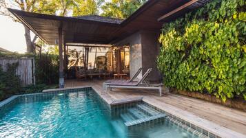 A swimming pool with wooden deck and lounge chairs, surrounded by lush greenery and a covered patio area.