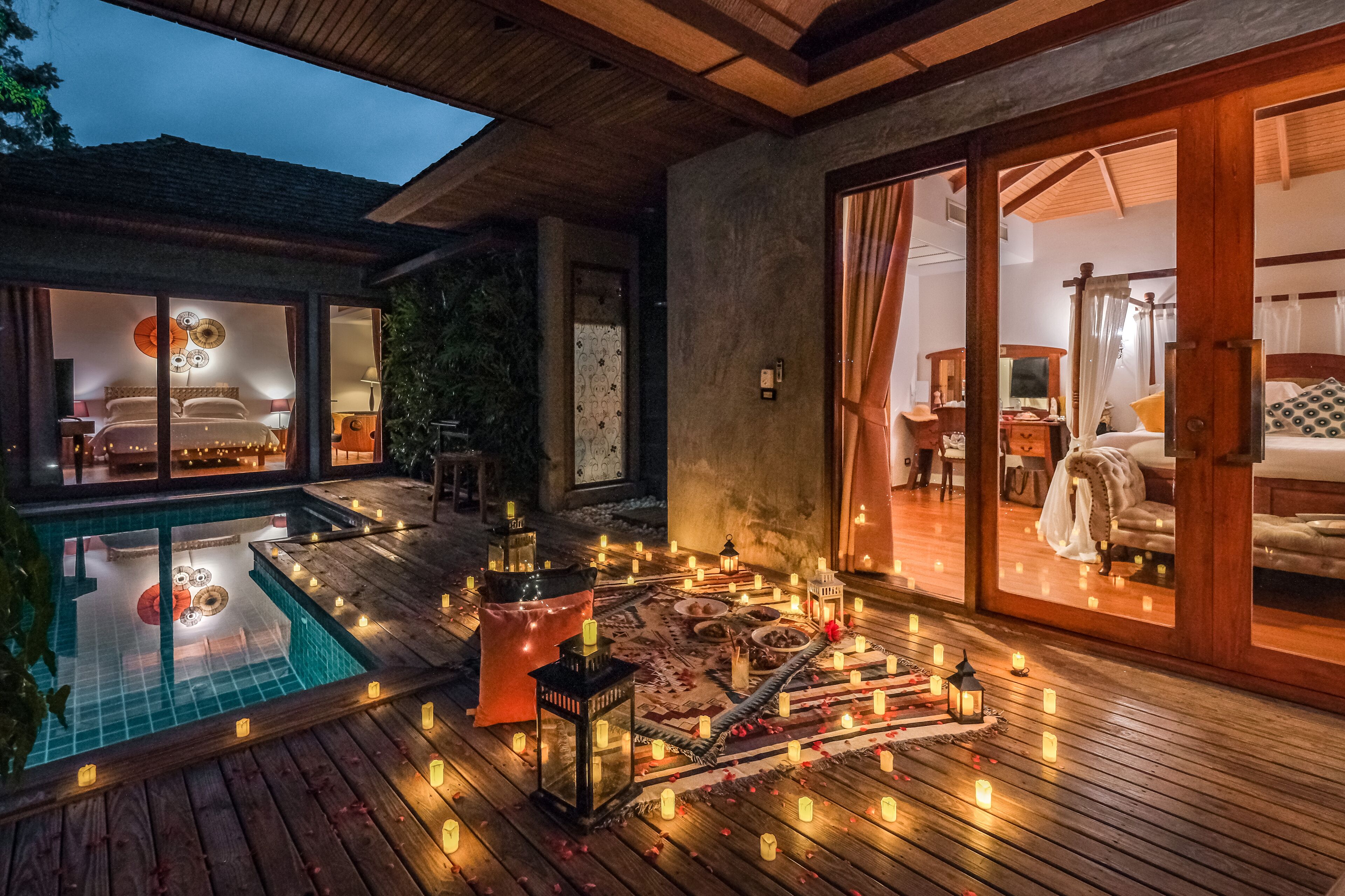 A well-lit outdoor area with a pool, wooden deck, and a cozy seating area with cushions and lanterns.