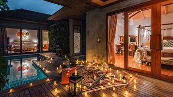 A well-lit outdoor area with a pool, wooden deck, and a cozy seating area with cushions and lanterns.