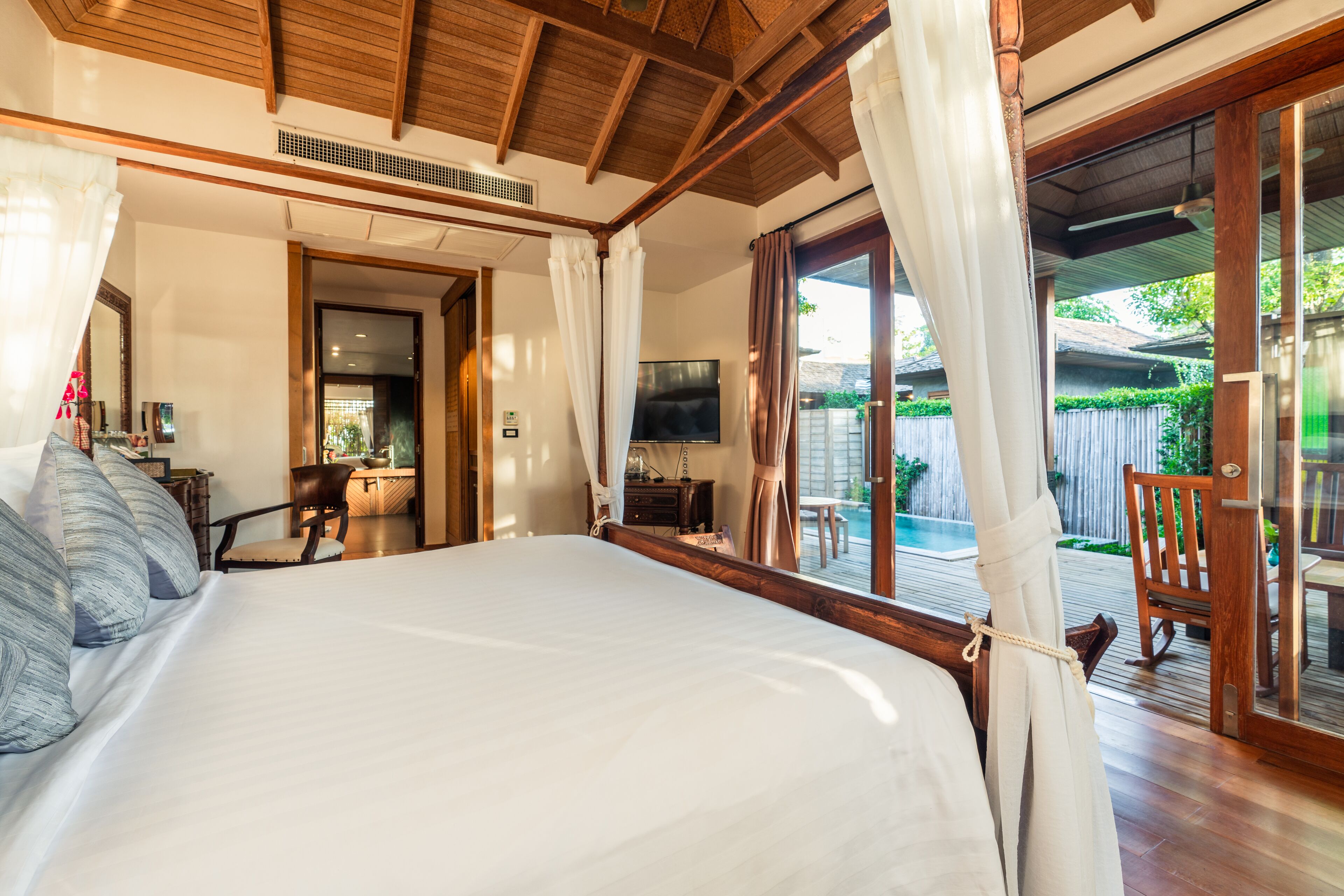 A bedroom with a large bed, wooden ceiling, and a view of a pool and outdoor seating area.