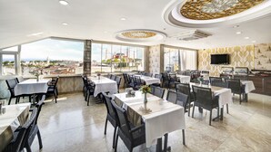 Un comedor amplio con varias mesas y sillas, ventanas grandes que ofrecen vista a un paisaje urbano y un diseño decorativo en el techo.