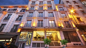 Un hotel de varios pisos con un diseño arquitectónico clásico, con balcones con barandillas de hierro forjado y una entrada bien iluminada.