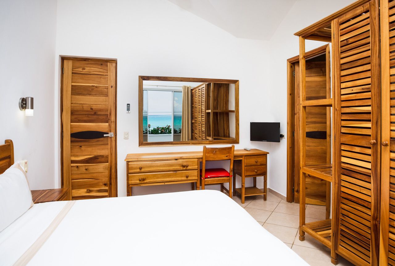Un dormitorio con un escritorio de madera, una silla, una cama, un televisor y un amplio ventanal con vista al mar.