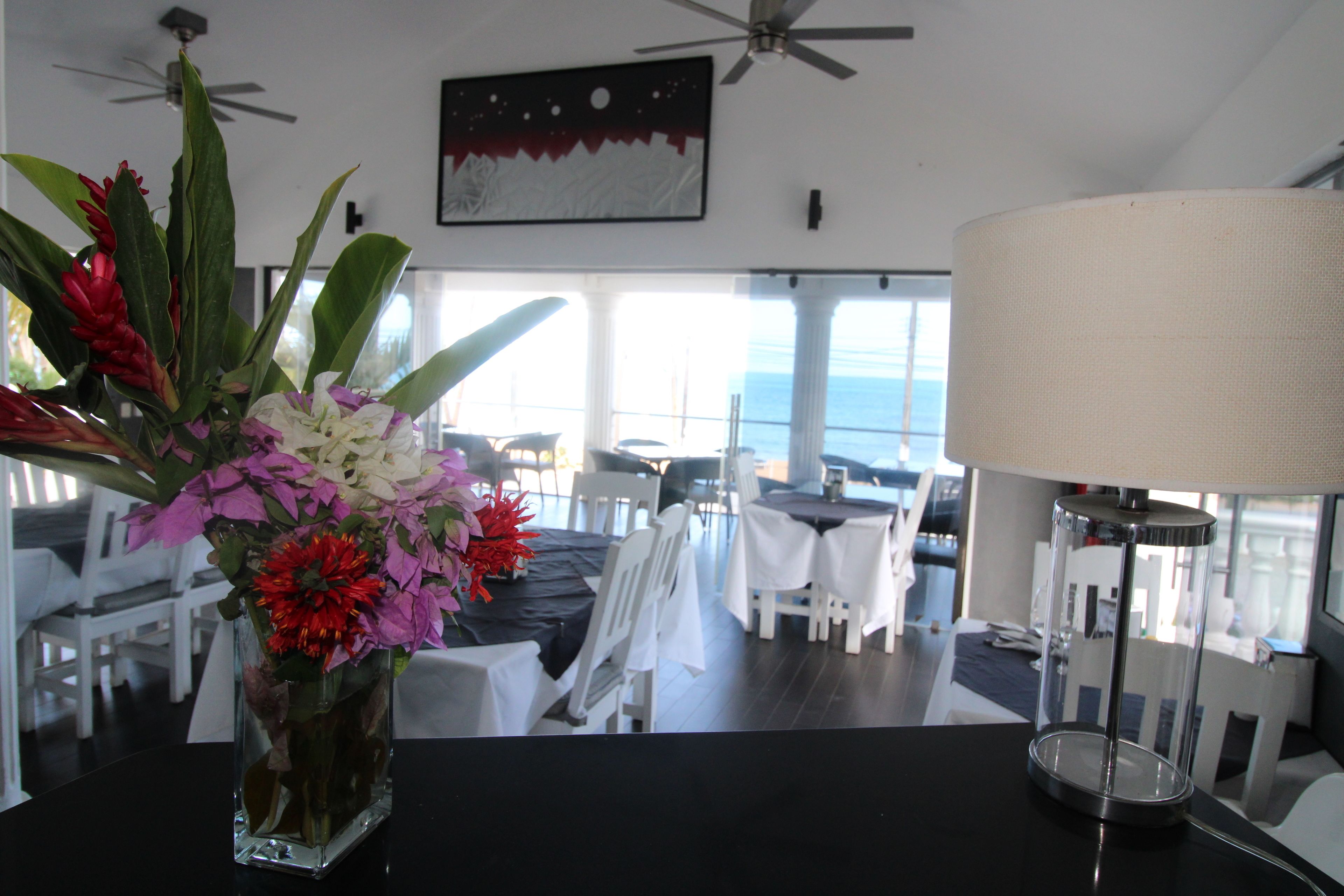 A dining area with a table, chairs, a vase of flowers, a ceiling fan, and a view of the ocean.