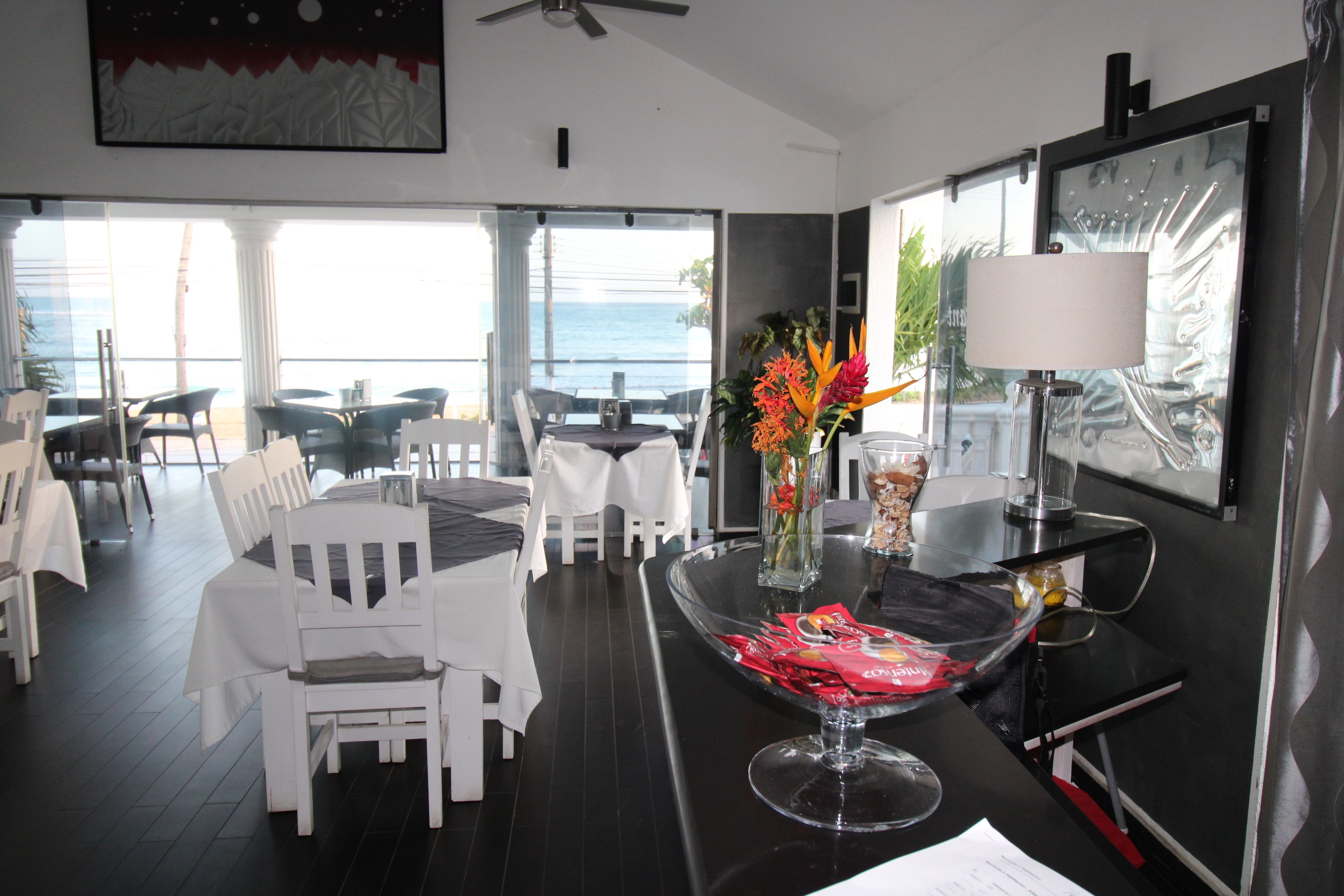 A modern dining area with white chairs and tables, a clear view of the ocean, and a decorative vase with flowers.