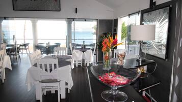 A modern dining area with white chairs and tables, a clear view of the ocean, and a decorative vase with flowers.