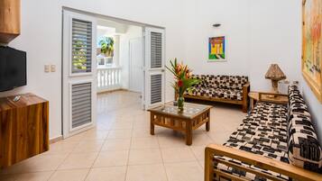 Amplia sala de estar con sofá, mesa de centro y mueble para televisión. Hay puertas abiertas que dan a un espacio exterior con vista a palmeras.