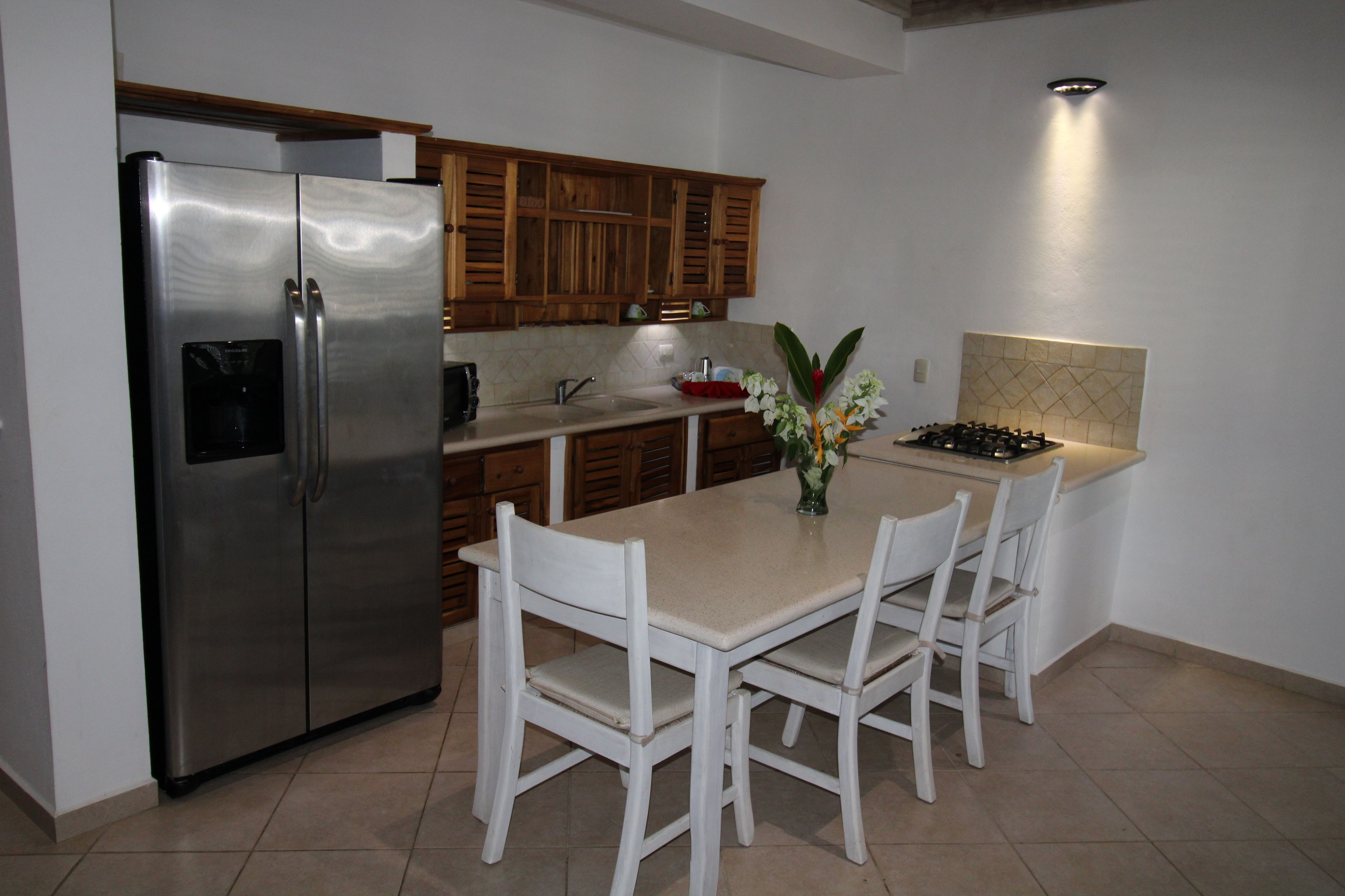 A kitchen with a stainless steel refrigerator, wooden cabinets, a white dining table with four chairs, and a vase with flowers.