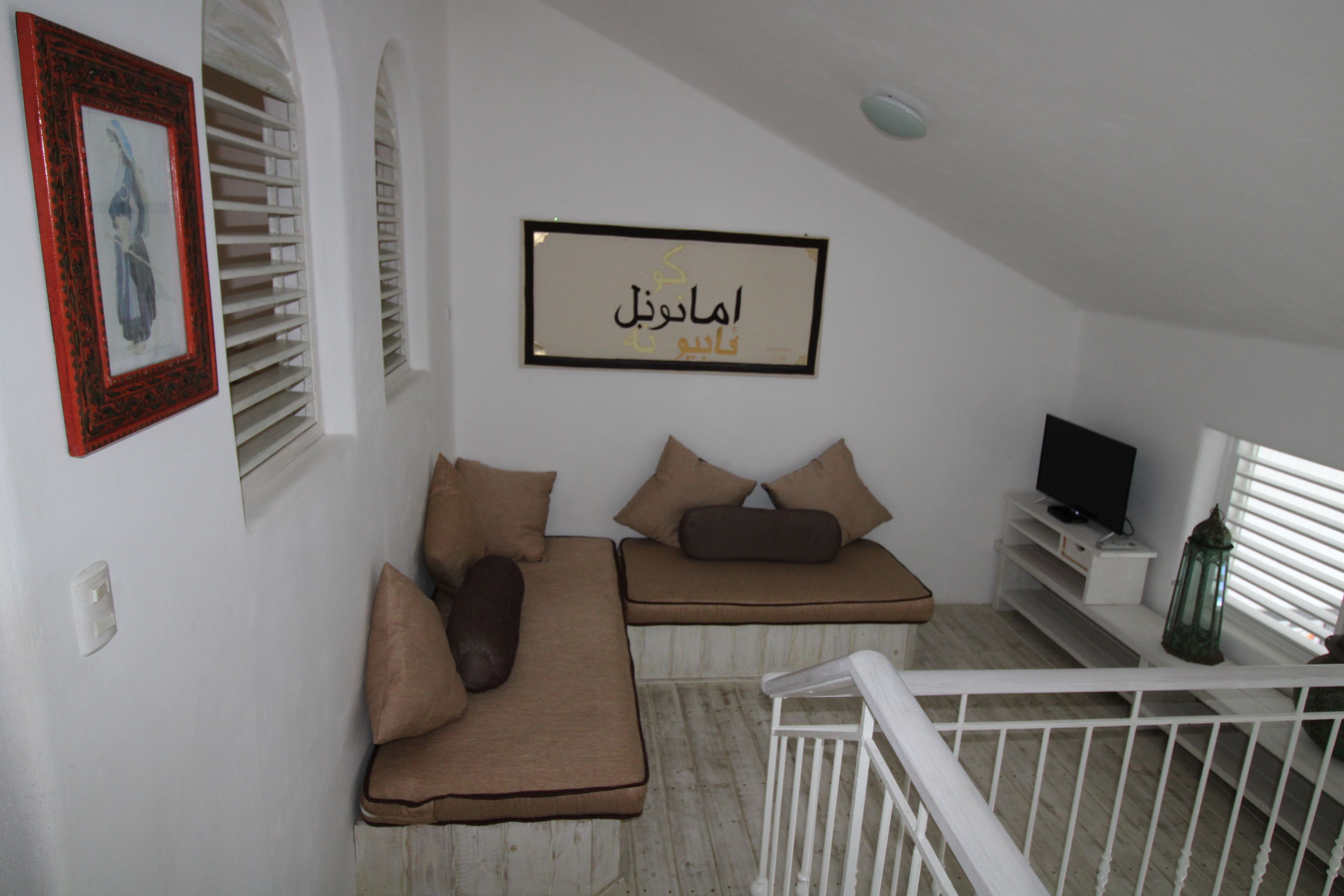A room with a white ceiling, a framed Arabic calligraphy on the wall, a white railing, and a television on a white stand.