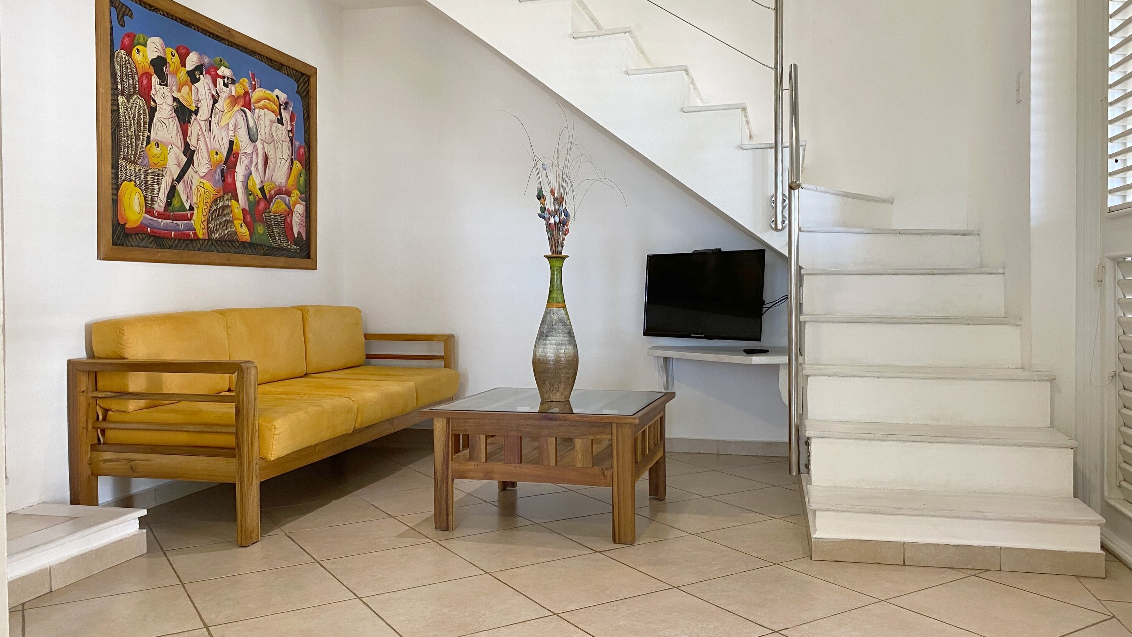 Un salon avec un canapé jaune, une table basse en bois, un tableau, un vase et un escalier.