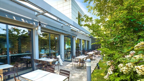 Un área de comedor al aire libre con mesas y sillas, amplias ventanas y un edificio moderno al fondo.