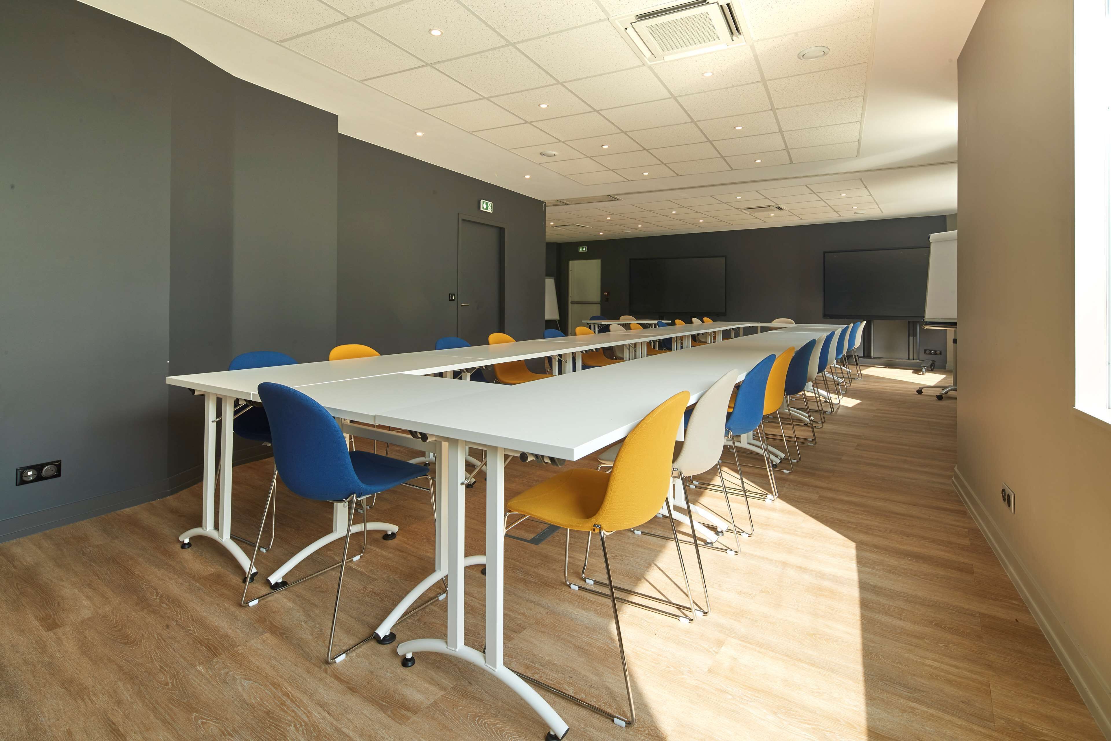 A conference room with a long table, blue and yellow chairs, a whiteboard, and a projector screen.